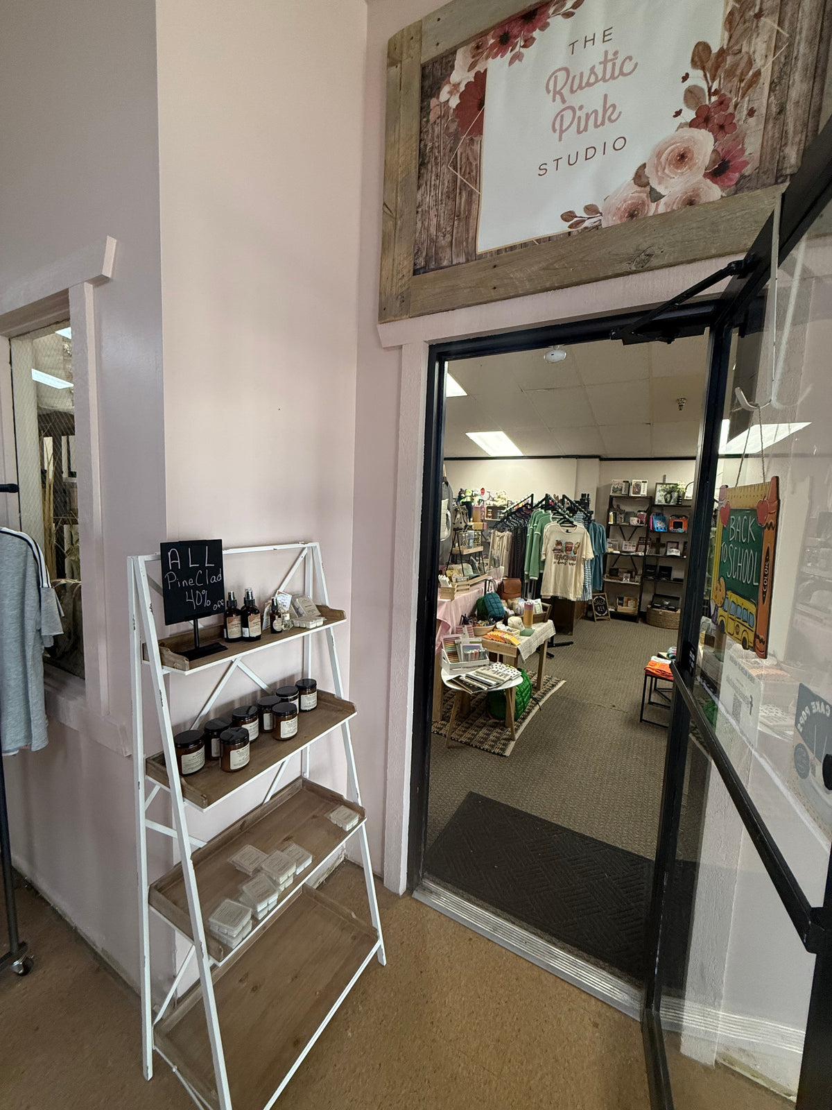 View from the entrance of 'The Rustic Pink Studio' looking into a boutique interior, with a white shelving unit holding small products on the left and clothing racks visible inside.